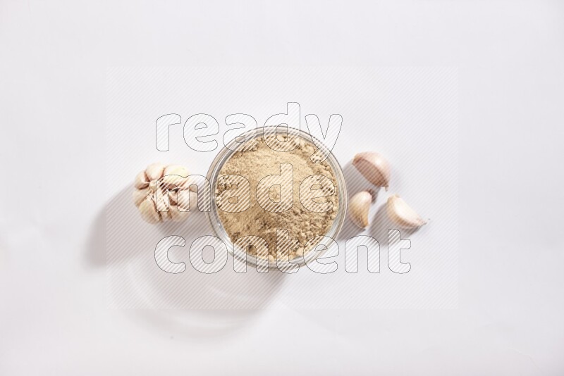 A glass bowl full of garlic powder with garlic bulb and some cloves beside it on a white flooring