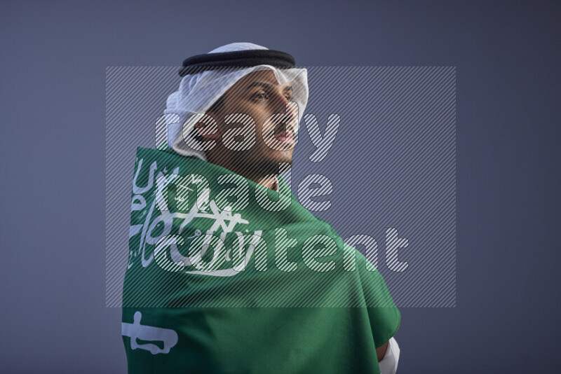 A close-up shot of Saudi man wearing thob and white shomag wrapping big Saudi flag on gray background