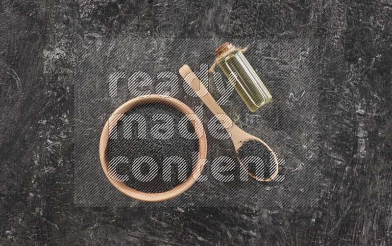 A wooden bowl and spoon full of black seeds and a bottle of black seeds oil on a textured black flooring