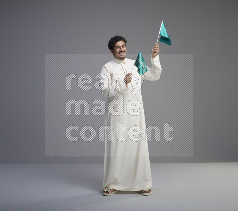 A Saudi man standing wearing thob holding small Saudi flag on gray background