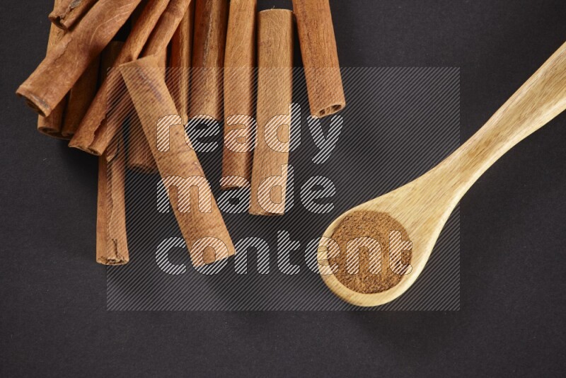 Cinnamon sticks stacked beside a wooden spoon full of cinnamon powder on black background