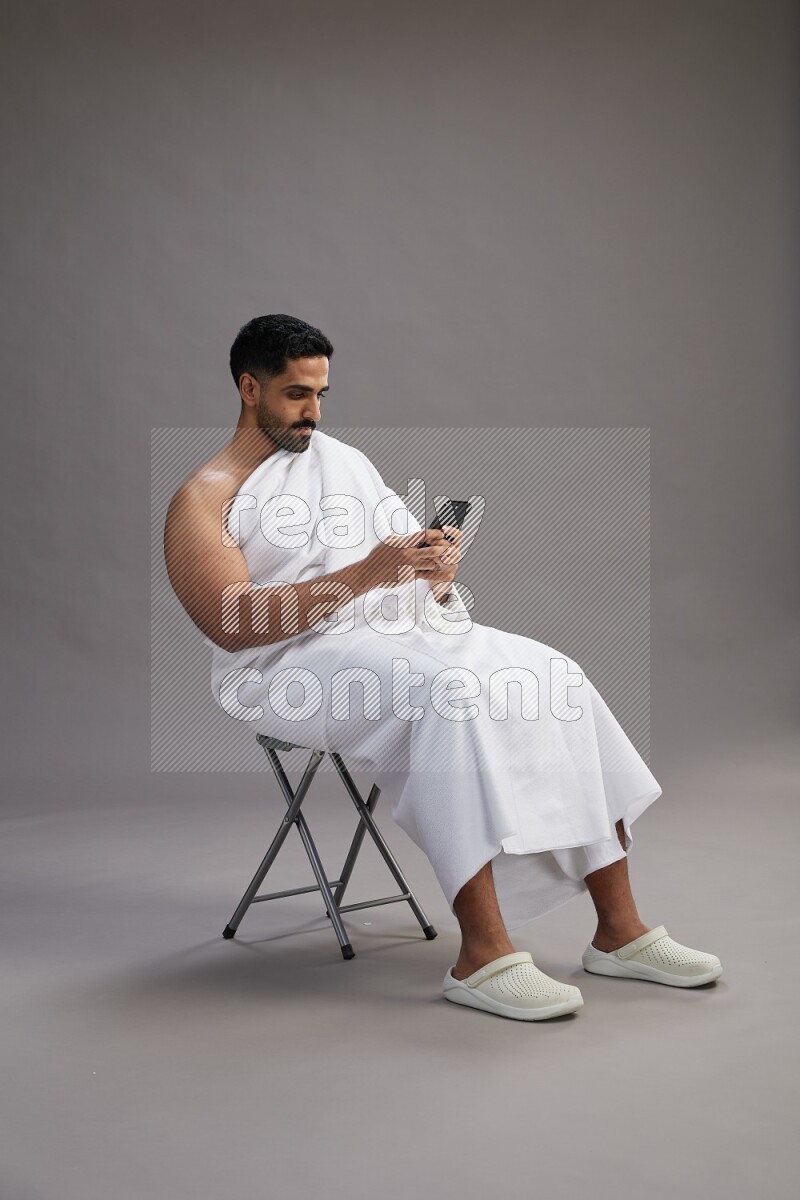 A man wearing Ehram sitting on chair texting on phone on gray background