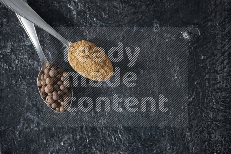 ملعقتان معدنيتان ممتلئتان ببودرة بهار الفلفل الحلو وحبوبه على خلفية سوداء