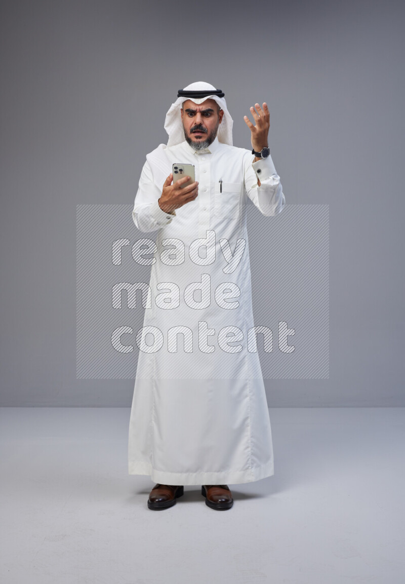 Saudi man Wearing Thob and white Shomag standing texting on phone on Gray background