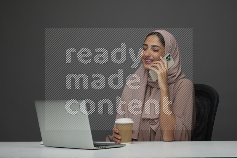 A Saudi woman Setting on her desk
 calling  on a Gray Background wearing Brown Abaya with Hijab
