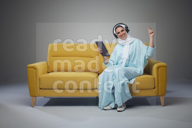 A Saudi woman wearing a light blue Abaya and a white head scarf sitting on a yellow sofa and holding an iPad while wearing headphone eye level on a grey background