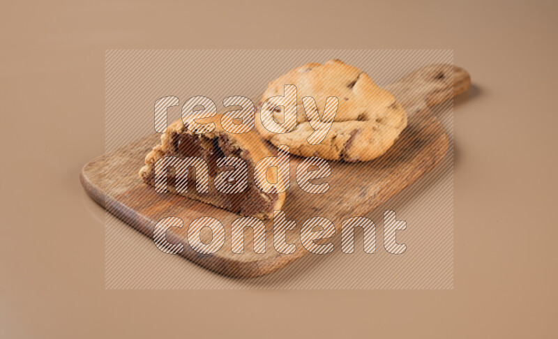 a chocolate chip cookie with another one cut in half on a wooden cutting board on a brown background