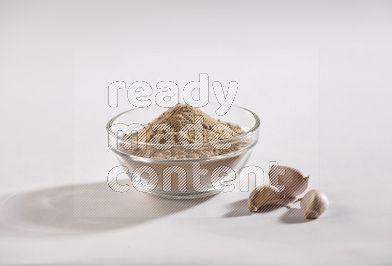 A glass bowl full of garlic powder with garlic bulb and some cloves beside it on a white flooring
