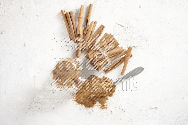 Flipped herbs glass jar full of cinnamon powder with a metal spoon full of powder and cinnamon sticks on a textured white background