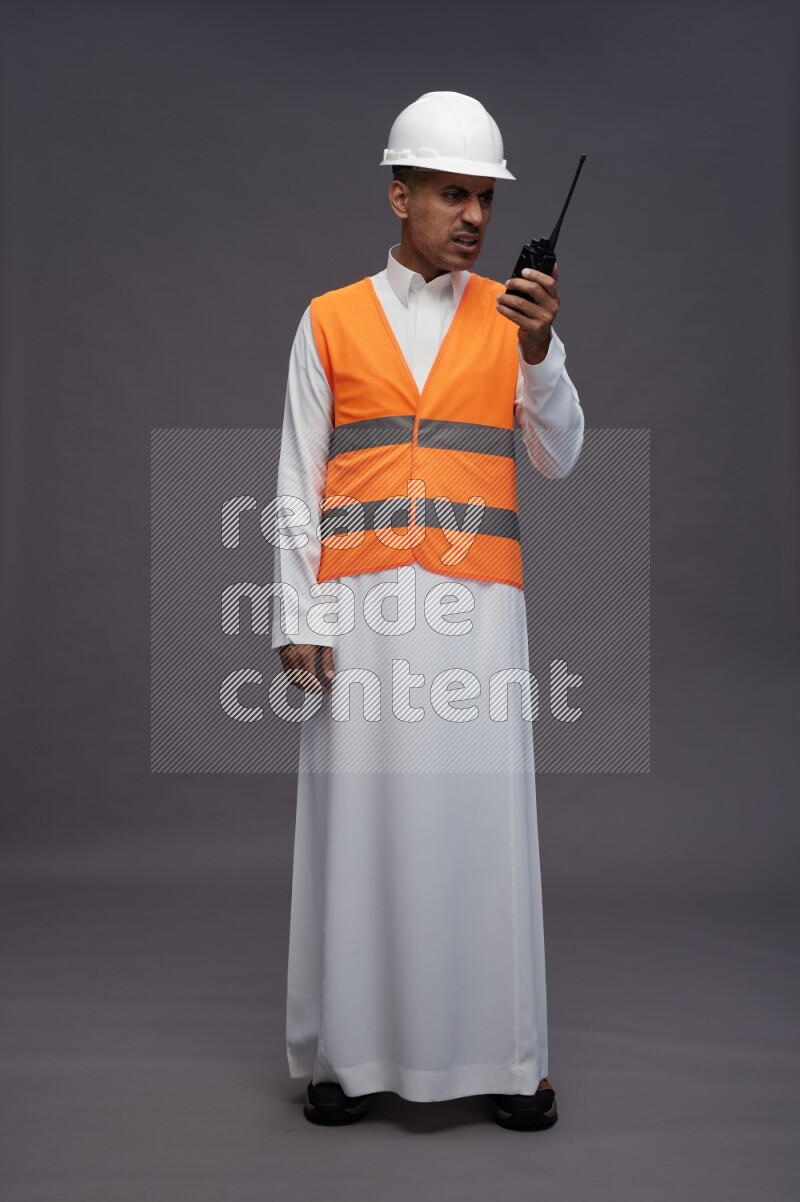 Saudi man wearing thob with engineer vest standing holding walkie-talkie on gray background