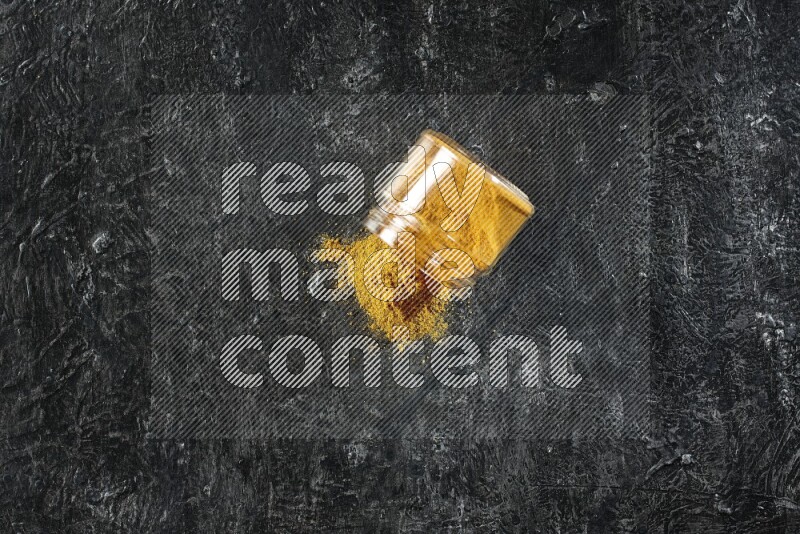 A flipped glass jar full of turmeric powder and powder spilled out of it on a textured black flooring