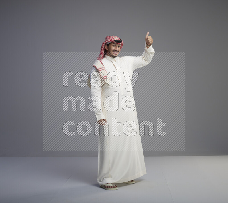 A Saudi man standing wearing thob and red shomag interacting with the camera on gray background