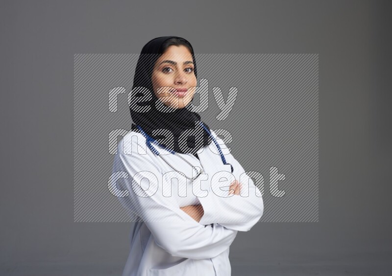 Saudi woman wearing lab coat with stethoscope standing with crossed arms on Gray background