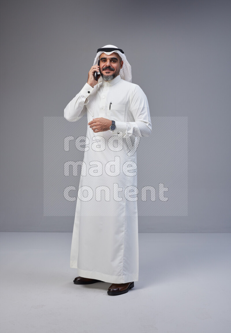 Saudi man Wearing Thob and white Shomag standing talking on phone on Gray background