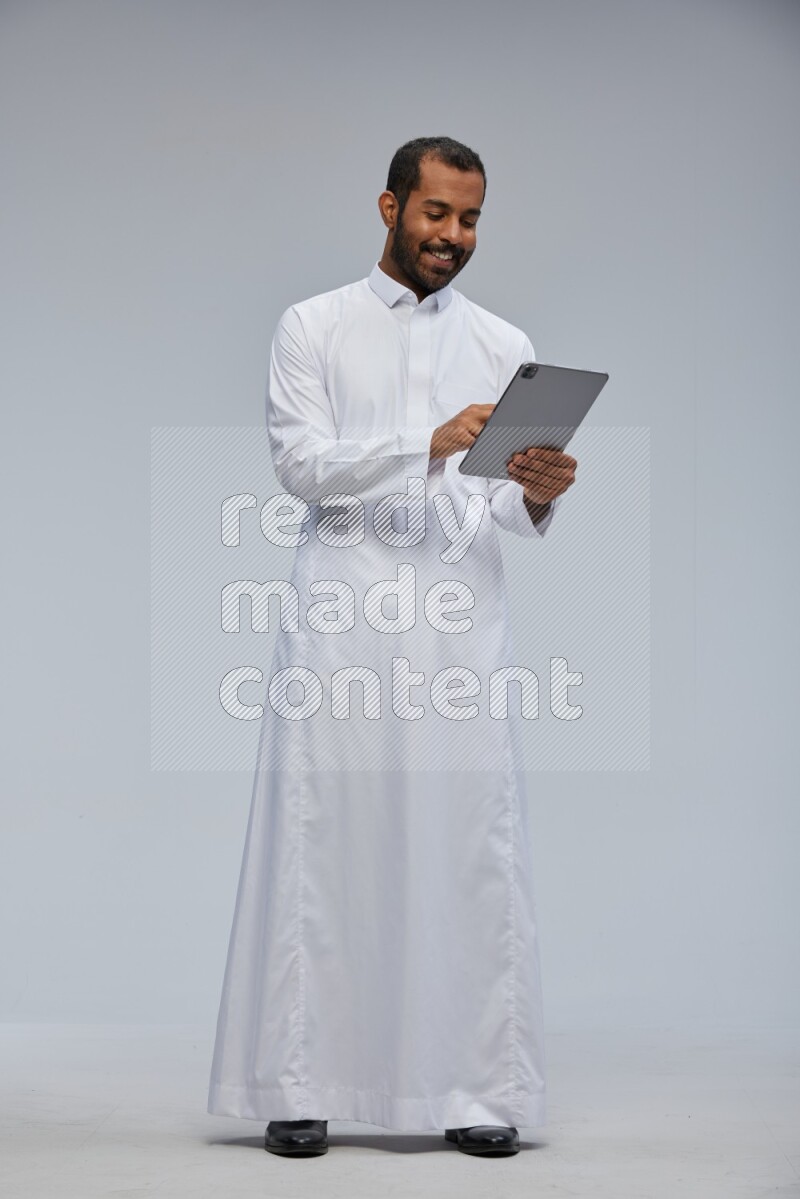 Saudi man wearing Thob standing working on tablet on Gray background