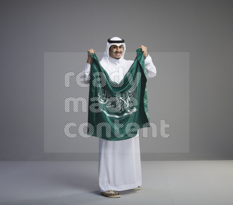 A Saudi man standing wearing thob and white shomag with face painting holding big Saudi flag on gray background