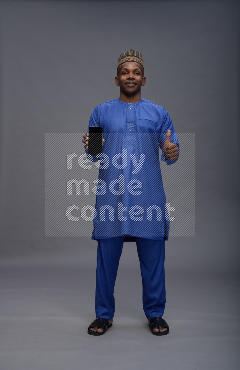 Man wearing Nigerian outfit standing showing phone to camera on gray background