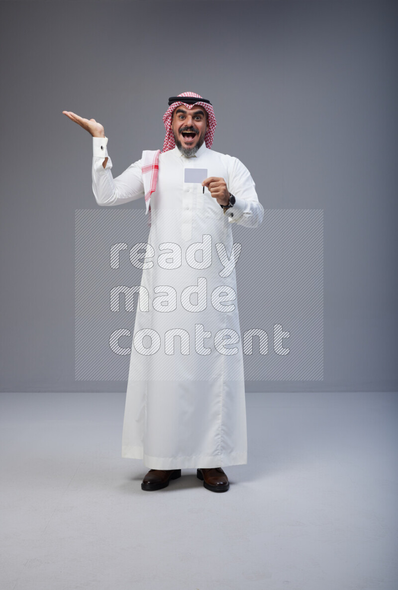 Saudi man Wearing Thob and red Shomag standing holding ATM card on Gray background