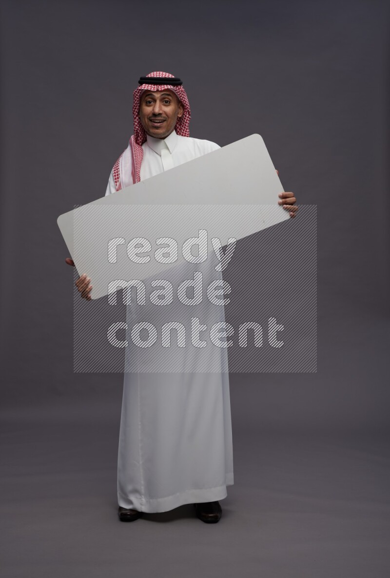 Saudi man wearing thob and shomag standing holding board on gray background