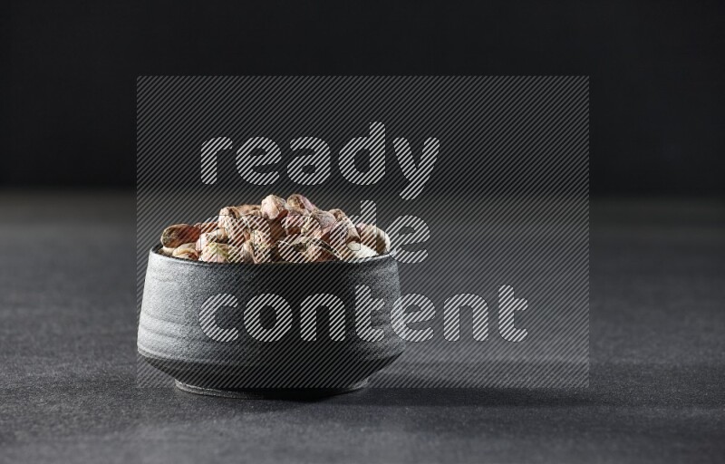 A black pottery bowl full of peeled pistachios on a black background in different angles