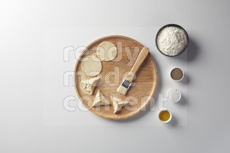 two closed sambosas and one open sambosa filled with cheese while flour, salt, black pepper and oil with oil brush aside in a wooden dish on a white background