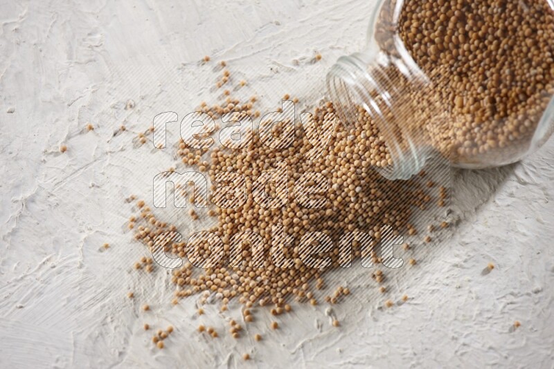 A glass spice jar full of mustard seeds and jar is flipped with fallen seeds on a textured white flooring