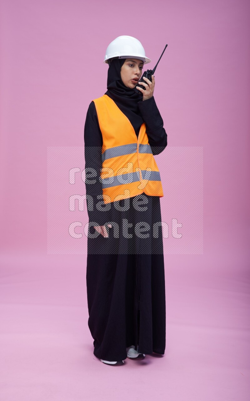 Saudi woman wearing Abaya with engineer vest and helmet standing holding walkie-talkie on pink background