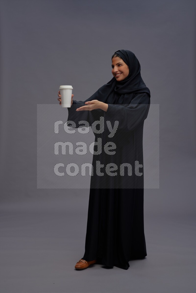 Saudi woman wearing Abaya standing holding paper cup on gray background