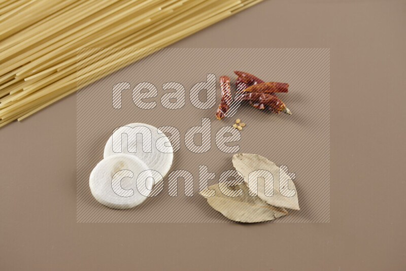 Raw pasta with different ingredients such as cherry tomatoes, garlic, onions, red chilis, black pepper, white pepper, bay laurel leaves, rosemary and cardamom on beige background