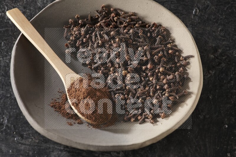 A Pottery plate full of cloves and a wooden spoon full of cloves powder on it on a textured black background