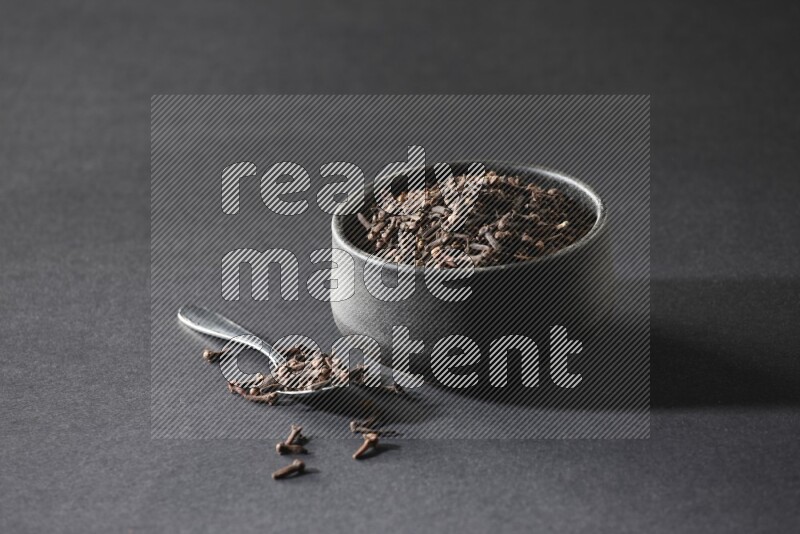 A black pottery bowl full of cloves and a metal spoon next to it on a black flooring