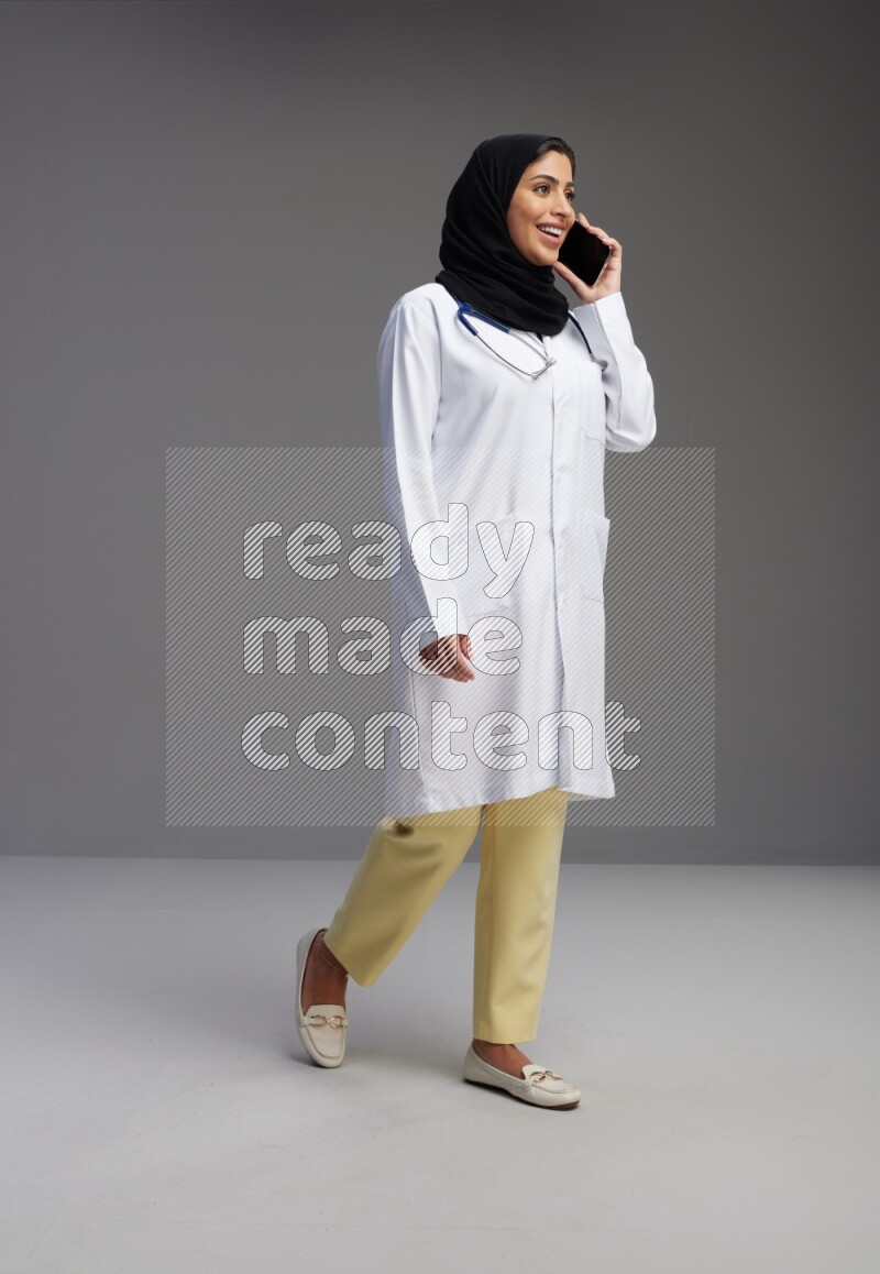 Saudi woman wearing lab coat with stethoscope standing talking on phone on Gray background