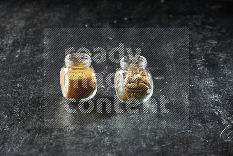 2 glass spice jars full of turmeric powder and dried whole fingers on textured black flooring
