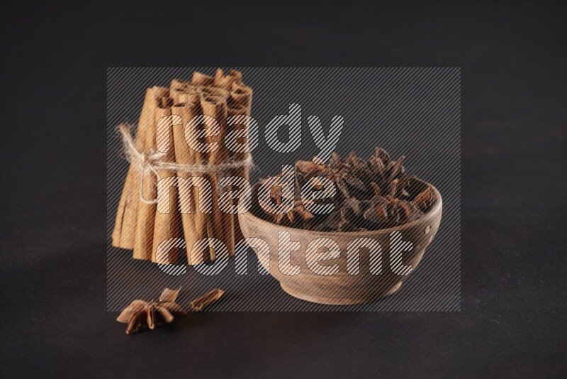 A stacked and bounded cinnamon sticks and a wooden bowl full of star anise on a black background