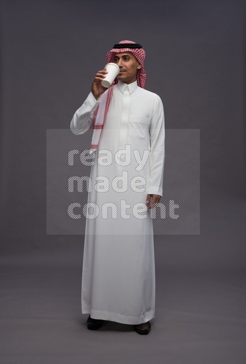 Saudi man wearing thob and shomag standing holding paper cup on gray background