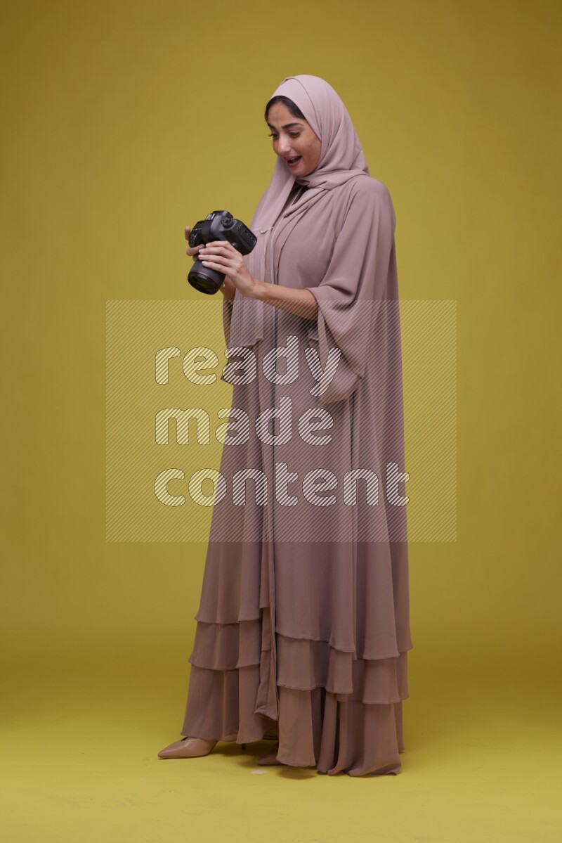 A woman Holding a Camera on a Yellow Background wearing Brown Abaya with Hijab