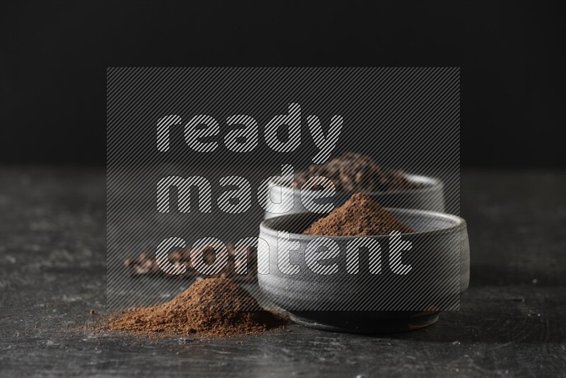 2 Black pottery bowls full of cloves and the other full of cloves powder on textured black flooring