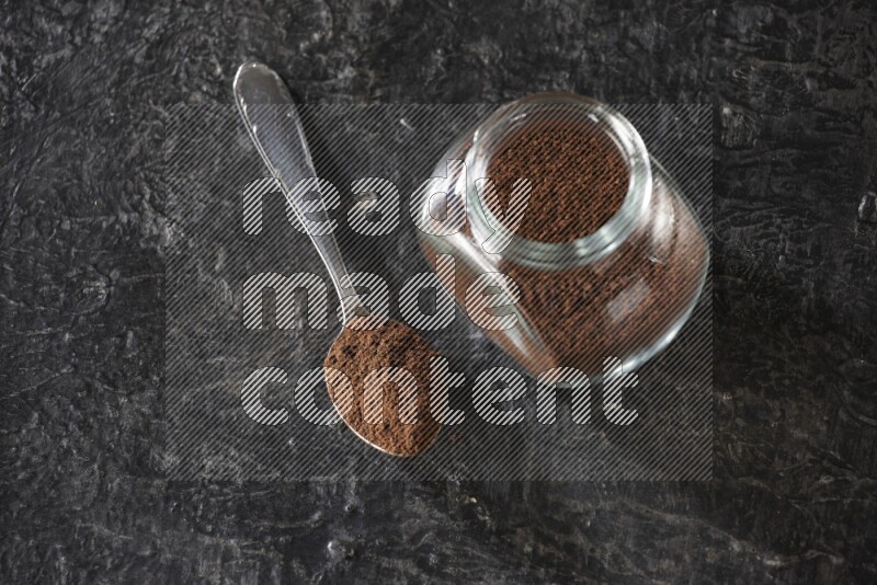 A glass spice jar and a metal spoon full of cloves powder on textured black flooring