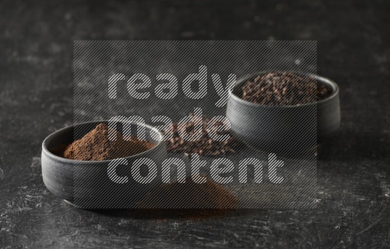 2 Black pottery bowls full of cloves and the other full of cloves powder on textured black flooring