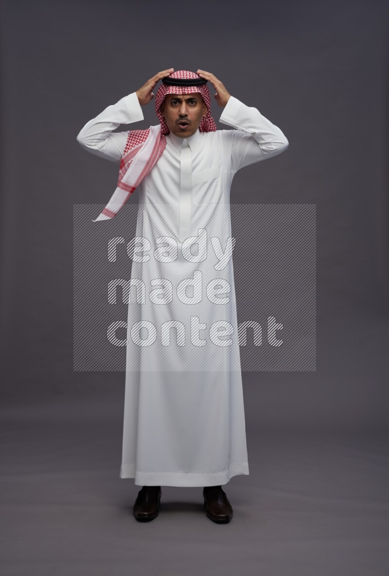 Saudi man wearing thob and shomag standing hands behind head on gray background