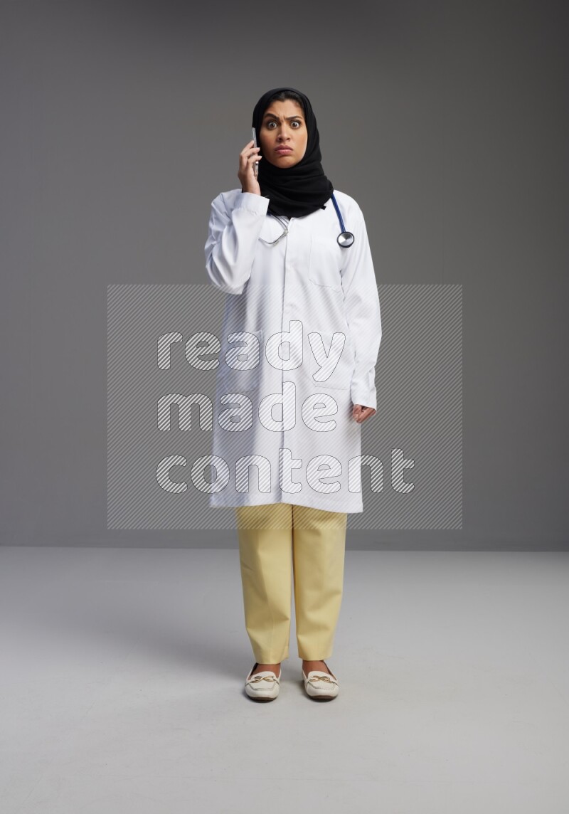 Saudi woman wearing lab coat with stethoscope standing talking on phone on Gray background