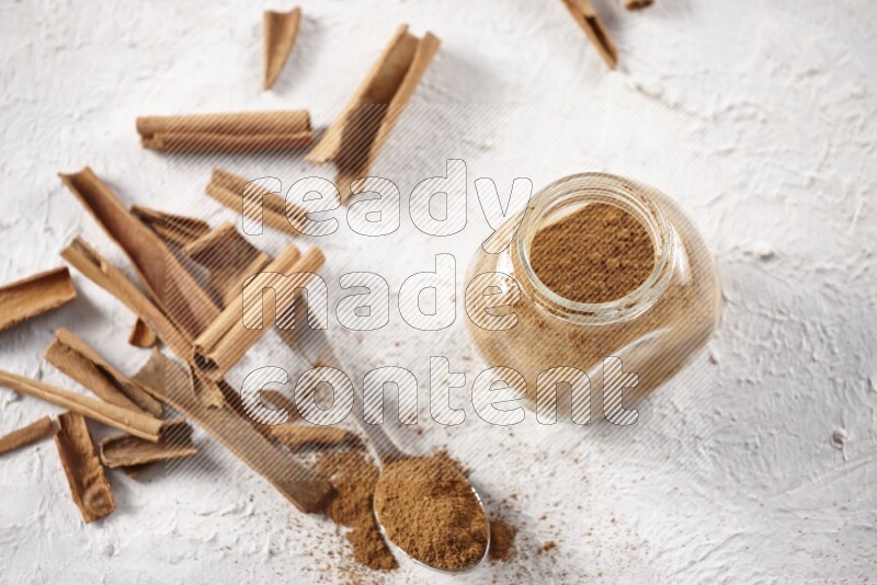 Herbal glass jar full cinnamon powder and a metal spoon surrounded by cinnamon sticks on a white background