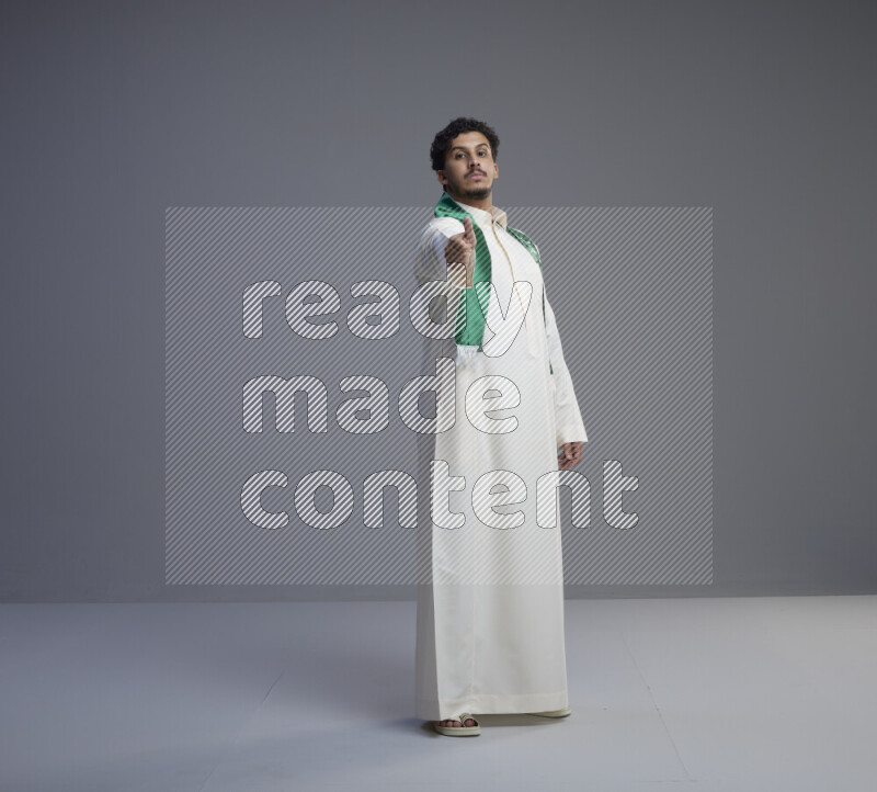 A Saudi man standing wearing thob and Saudi flag scarf on gray background