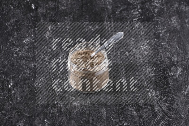 بودرة الفلفل الأسود في وعاء زجاجي مع ملعقة معدنية علي خلفية سوداء