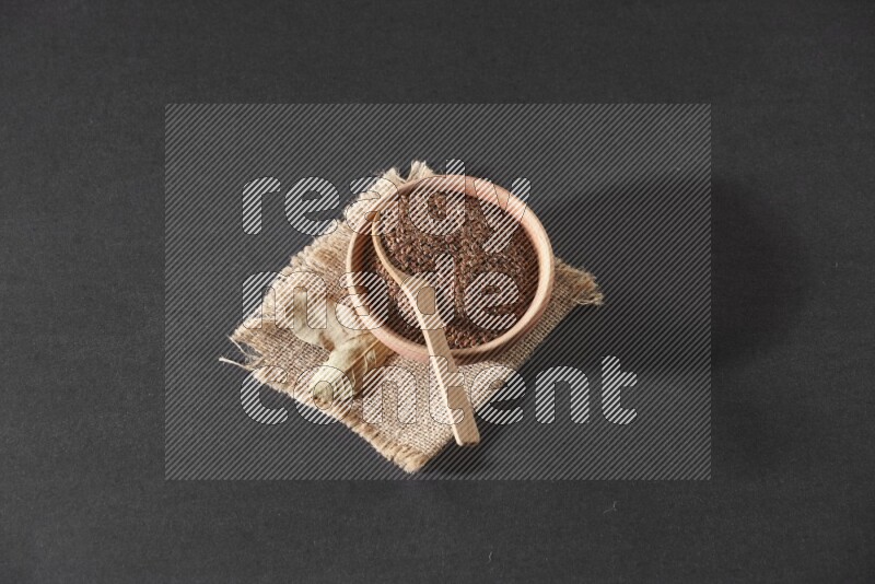A wooden bowl full of flaxseeds with wooden spoon full of the seeds on it on burlap fabric on a black flooring
