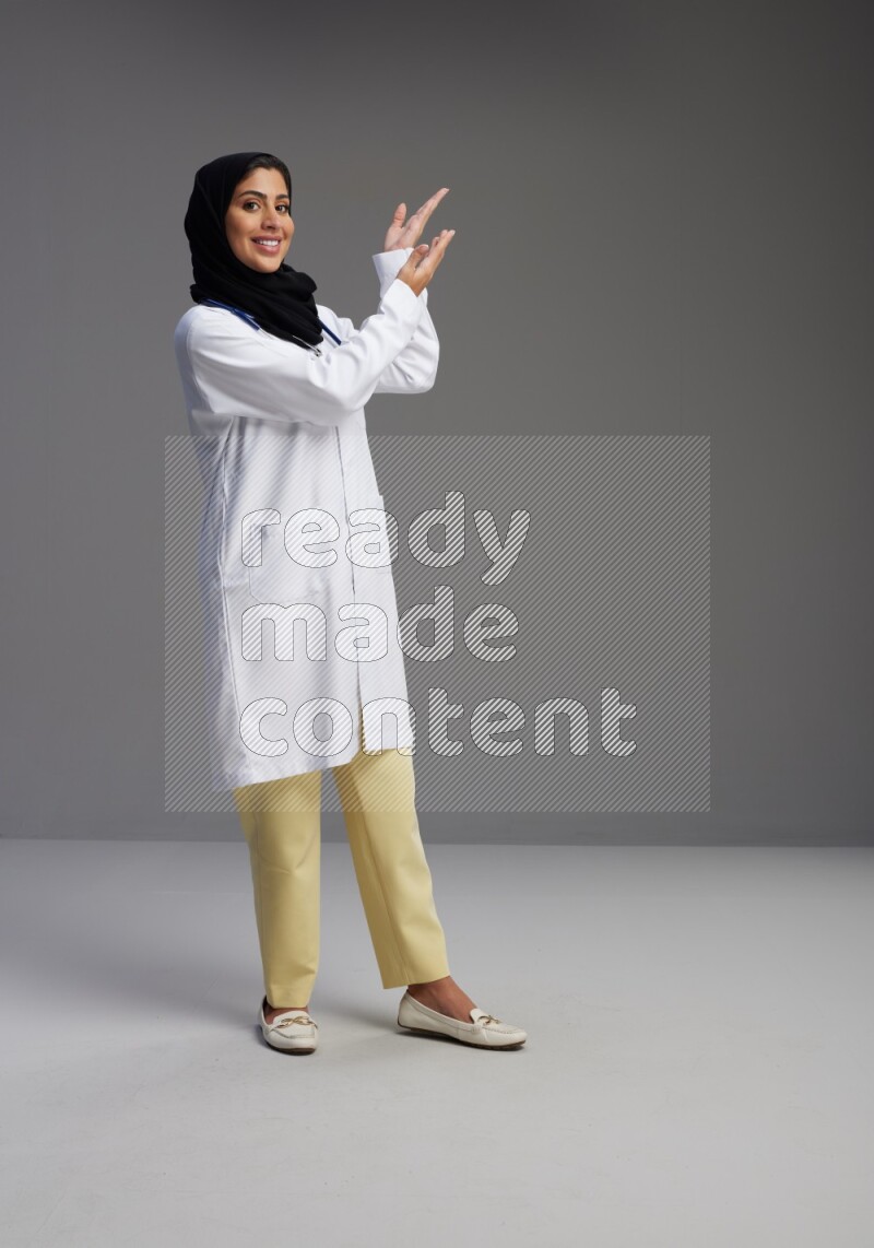 Saudi woman wearing lab coat with stethoscope standing interacting with the camera on Gray background