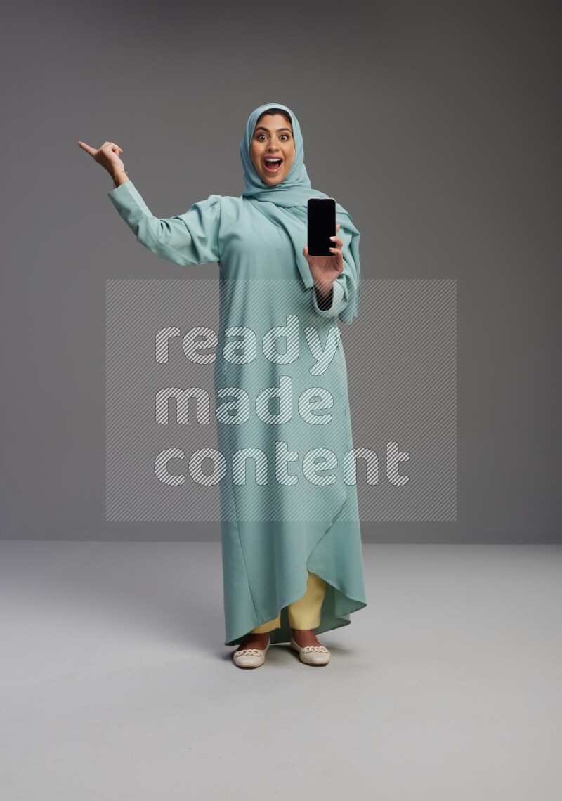 Saudi Woman wearing Abaya standing showing phone to camera on Gray background