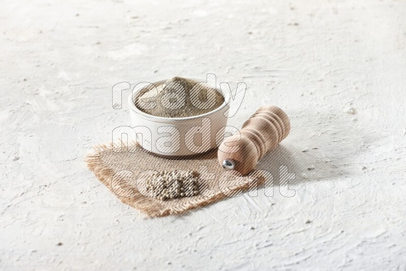 White pottery bowl full of white pepper powder set on a burlap piece of fabric with pepper beads and wooden pepper grinder on textured white flooring