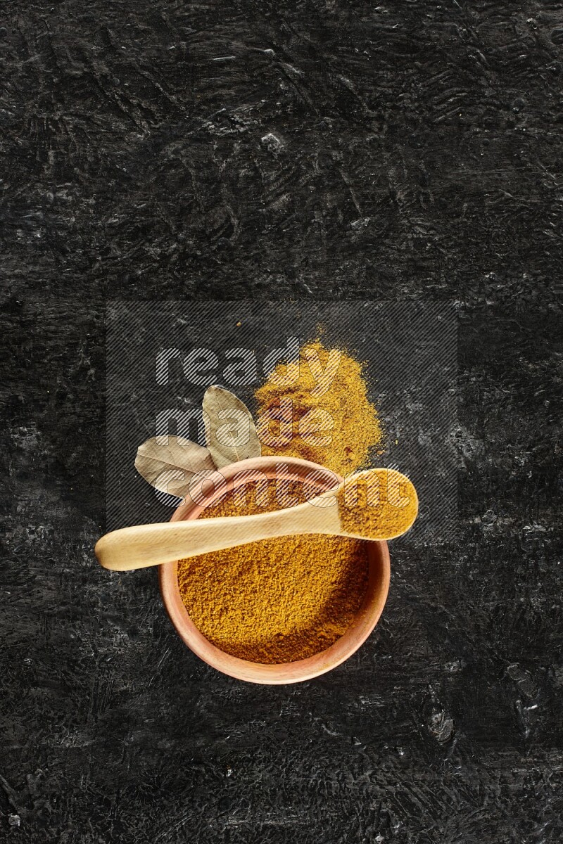A wooden bowl and a wooden spoon full of turmeric powder on textured black flooring