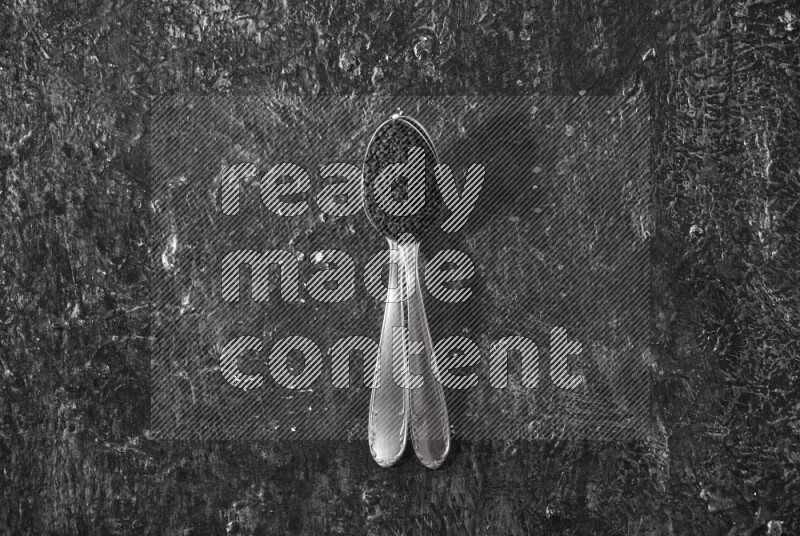 2 metal spoons full of black seeds on a textured black flooring